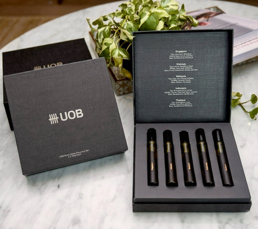 Open Black textured gift box displaying a UOB Room Spray Discovery Set with five scent vials and regional fragrance notes listed inside, set on a marble table with green foliage.